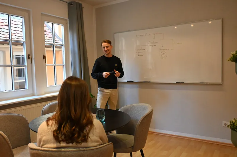 Marvin Gräber erklärt an einem Whiteboard, eine Zuhörerin sitzt an einem runden Tisch davor.